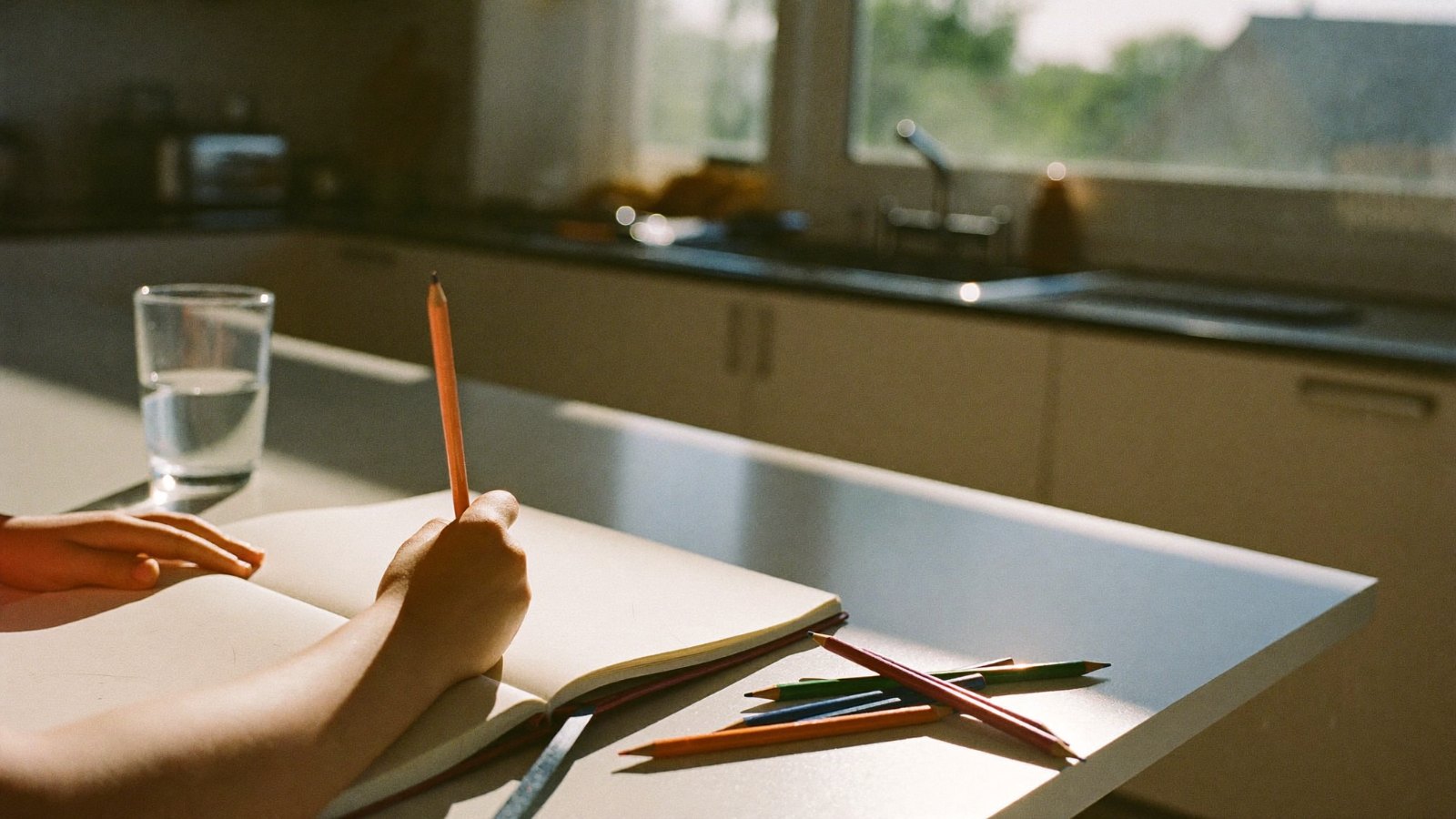 Child writing in a notebook at a kitchen table in morning light, coloured pencils beside