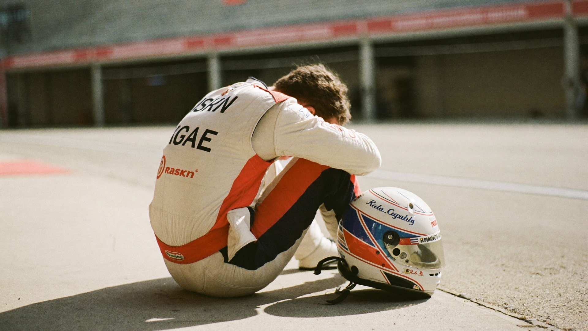 A racing driver in full race suit sits alone on the pit wall in quiet reflection before a race