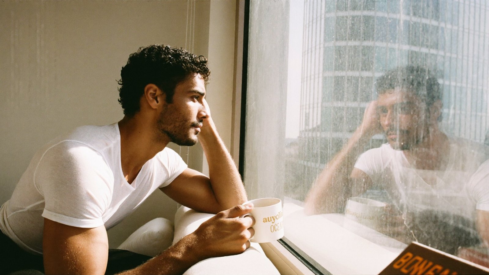 Man sitting by a window in morning light holding a mug of tea, looking out
